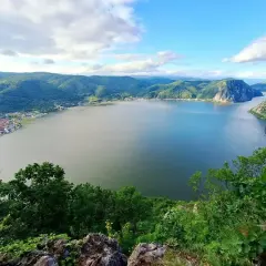 Szeroka rzeka Dunaj otoczona zielonymi wzgórzami i skalistymi zboczami. Na brzegu widać zabudowania.
