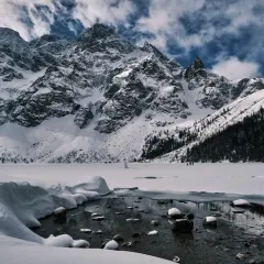 Morskie Oko zimową porą. Majestatyczne, ośnieżone szczyty Tatr górują nad zamarzniętym jeziorem. W oddali widać ludzi spacerujących po lodzie.