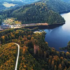 Malownicze jezioro Bystrzyckie otoczone jesiennym lasem. W oddali widoczny kompleks budynków nad brzegiem.