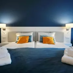 Przytulna sypialnia z dwoma łóżkami, pomarańczowymi poduszkami i białymi szlafrokami. Idealne miejsce na relaks, jak sugerują hotel hel opinie.