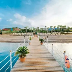 Drewniane molo prowadzi do plaży z rzędami leżaków i palm. W tle widać hotel, który zachęca do sprawdzenia mukarnas spa resort opinie.
