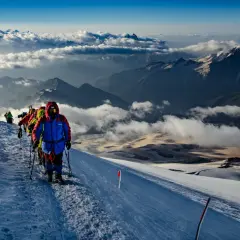 Grupa alpinistów wspina się po ośnieżonym zboczu, podziwiając majestatyczne góry z Elbrusem w tle. Chmury unoszą się poniżej, tworząc baśniowy krajobraz.