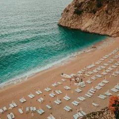 Plaża w Turcji, turkusowe morze, piaszczysty brzeg z rzędami leżaków i parasoli, skaliste wybrzeże.
