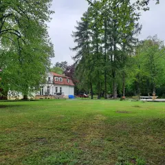 Biały dworek z czerwonym dachem otoczony zielenią. W oddali widać fontannę i drzewa. To urokliwa **gierłoż**.