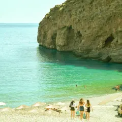 Piękna pogoda w Grecji w maju: turkusowa woda, skaliste klify i plaża z parasolami. Trzy dziewczyny idą w stronę morza.