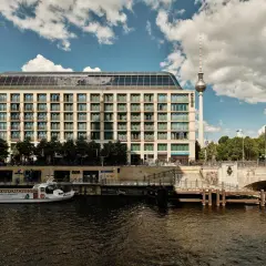 Nowoczesny budynek hotelu Radisson Blu Berlin nad rzeką Sprewą, z widoczną w tle wieżą telewizyjną.