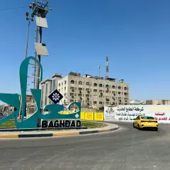 Wielki napis "BAGHDAD" i stylizowany napis arabski na tle budynków i nieba. Stolica Iraku wita podróżnych.