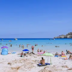 Słoneczny dzień na plaży Cala Agulla. Ludzie kąpią się w turkusowej wodzie, opalają na piasku i odpoczywają pod parasolami.