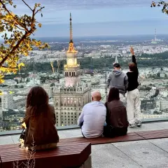 Ludzie podziwiają panoramę Warszawy z Pałacem Kultury i Nauki. Widać wiele warszawskich wież i budynków.