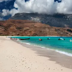Piękne wakacje na Sokotrze: piaszczysta plaża, turkusowe morze i łodzie kołyszące się na falach. W tle skaliste wzgórza i chmury.