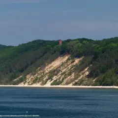 Zielone wzgórza porośnięte lasem schodzą ku morzu. Na szczycie wzniesienia, na tle błękitnego nieba, stoi ceglana wieża. Pomiędzy lasem a wodą rozciąga się piaszczysta plaża, tworząc białą górę.