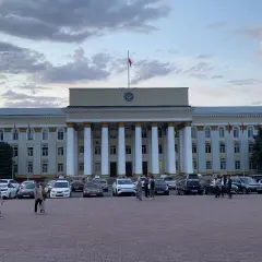 Plac przed budynkiem rządowym w Biszkeku, stolicy Kirgistanu. W tle monumentalna fasada z kolumnami, na dachu flaga. Na placu zaparkowane samochody i spacerujący ludzie.