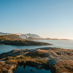 Most znany z pocztówek z fiordów norweskich, łukowato wznoszący się nad wodą, z górami w tle.