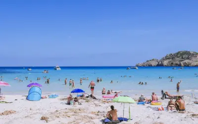 Słoneczny dzień na plaży Cala Agulla. Ludzie kąpią się w turkusowej wodzie, opalają na piasku i odpoczywają pod parasolami.