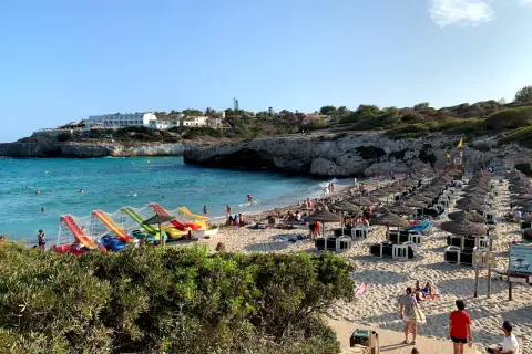 Słoneczna plaża Cala Domingos tętni życiem. Woda kusi turkusowym blaskiem, a piasek ugina się pod ciężarem leżaków i parasoli. Dzieci bawią się na kolorowych zjeżdżalniach.
