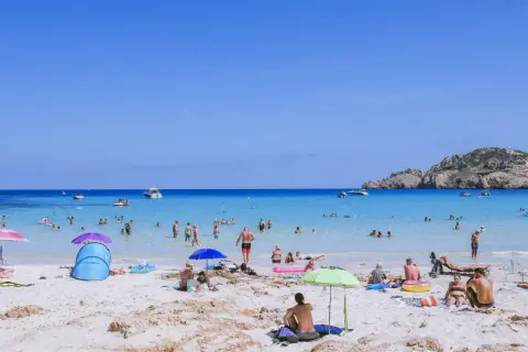 Słoneczny dzień na plaży Cala Agulla. Ludzie kąpią się w turkusowej wodzie, opalają na piasku i odpoczywają pod parasolami.