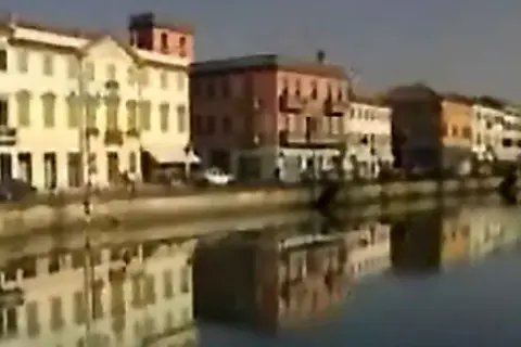 Nadbrzeże z kolorowymi kamienicami odbija się w spokojnej wodzie. W oddali widać fragmenty budynków nad rzeką.