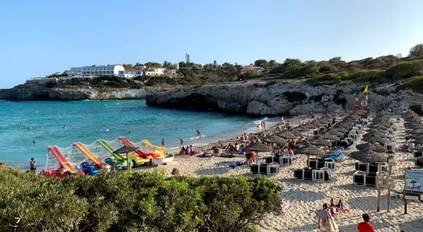 Słoneczna plaża Cala Domingos tętni życiem. Woda kusi turkusowym blaskiem, a piasek ugina się pod ciężarem leżaków i parasoli. Dzieci bawią się na kolorowych zjeżdżalniach.