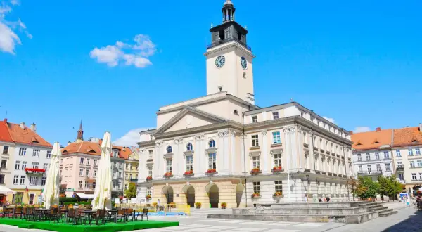 Rynek w mieście nad Prosną. Ratusz z wieżą zegarową góruje nad placem, gdzie stoją stoliki kawiarniane.