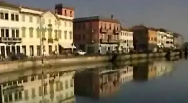 Nadbrzeże z kolorowymi kamienicami odbija się w spokojnej wodzie. W oddali widać fragmenty budynków nad rzeką.