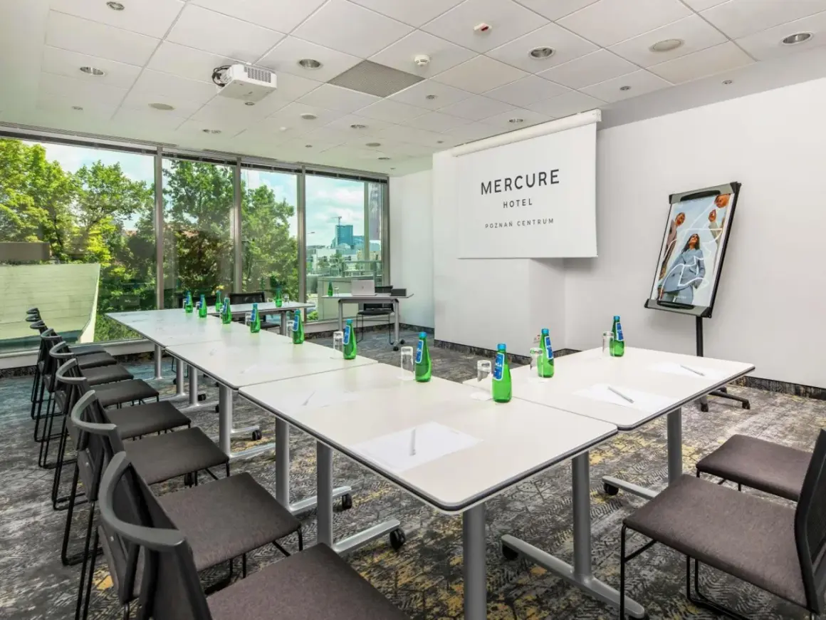 Sala konferencyjna w hotelu Mercure Poznań Centrum. Długi stół, krzesła, projektor i logo hotelu.