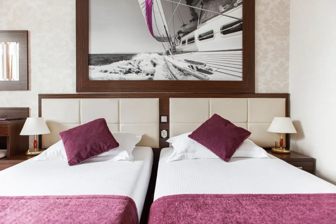 Dwie wygodne łóżka w pokoju hotelowym, nad nimi obraz z żaglówką. Idealne miejsce na wypoczynek, jak sugerują primavera jastrzębia góra opinie.