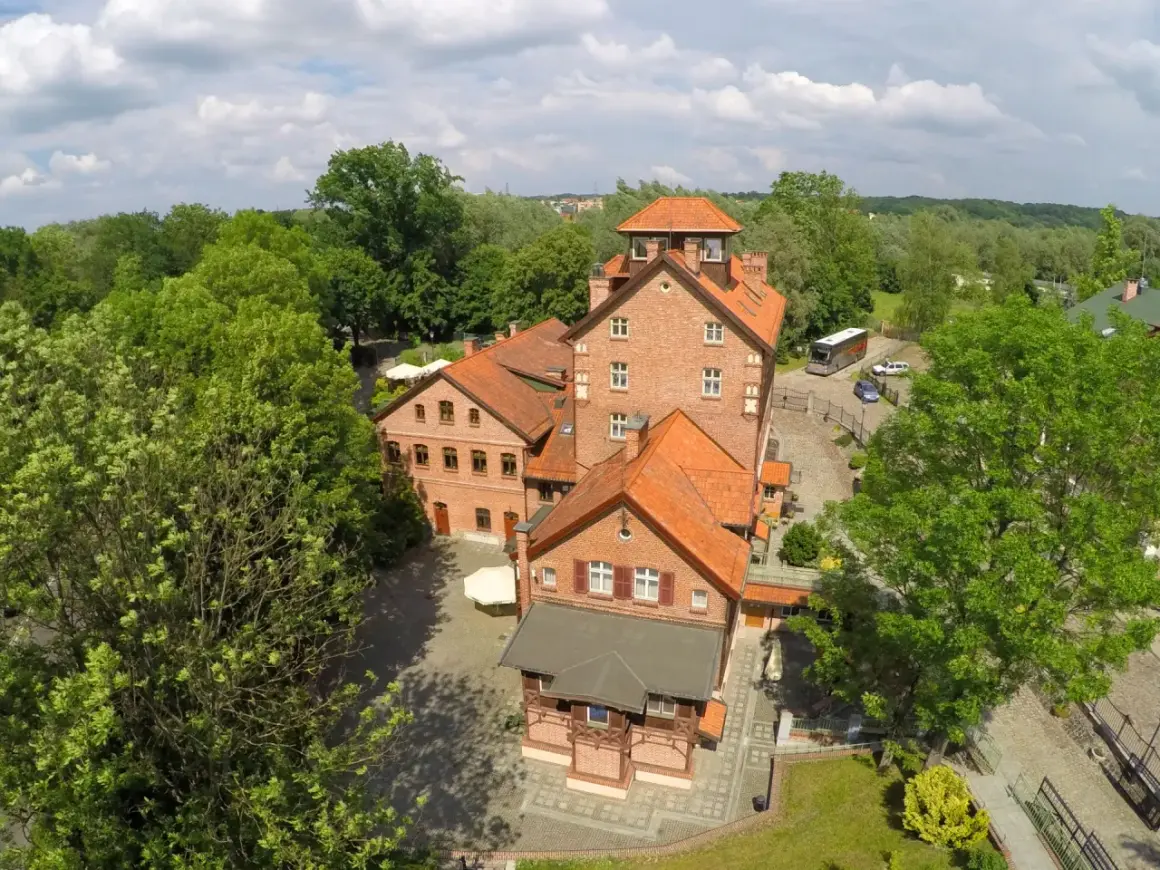 Hotel Młyn Elbląg, ceglane budynki z czerwonymi dachami, otoczone zielenią.