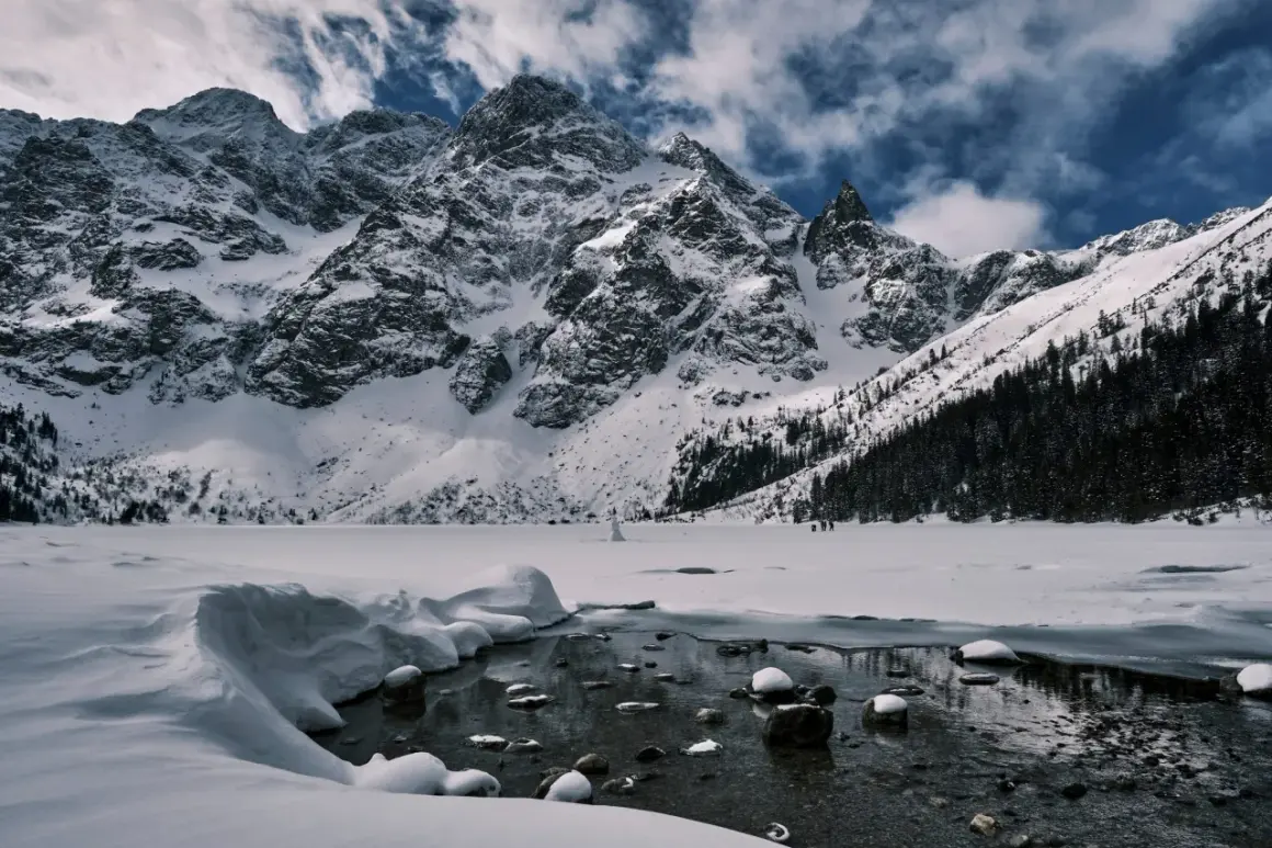 Morskie Oko zimową porą. Majestatyczne, ośnieżone szczyty Tatr górują nad zamarzniętym jeziorem. W oddali widać ludzi spacerujących po lodzie.