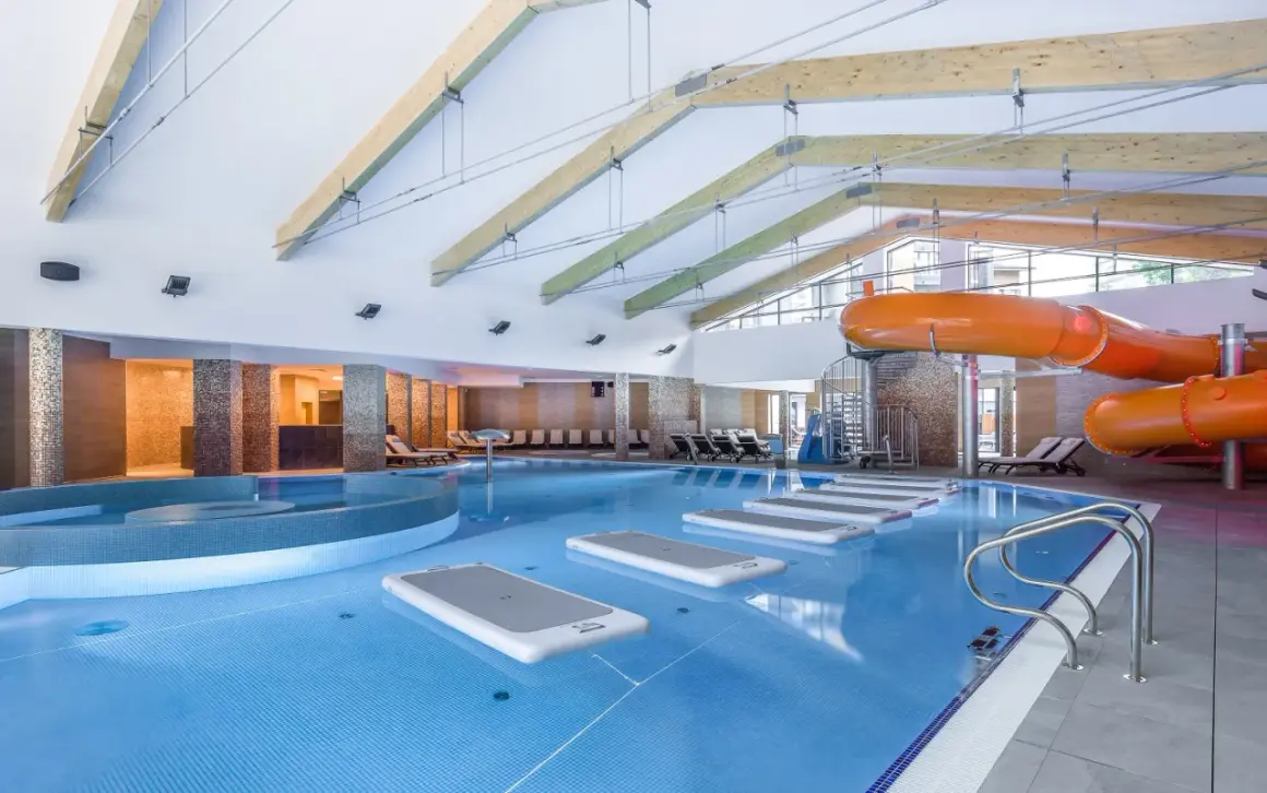Wnętrze hotelu z aquaparkiem w górach. Basen z pomarańczowymi zjeżdżalniami, leżakami i dmuchanymi materacami.