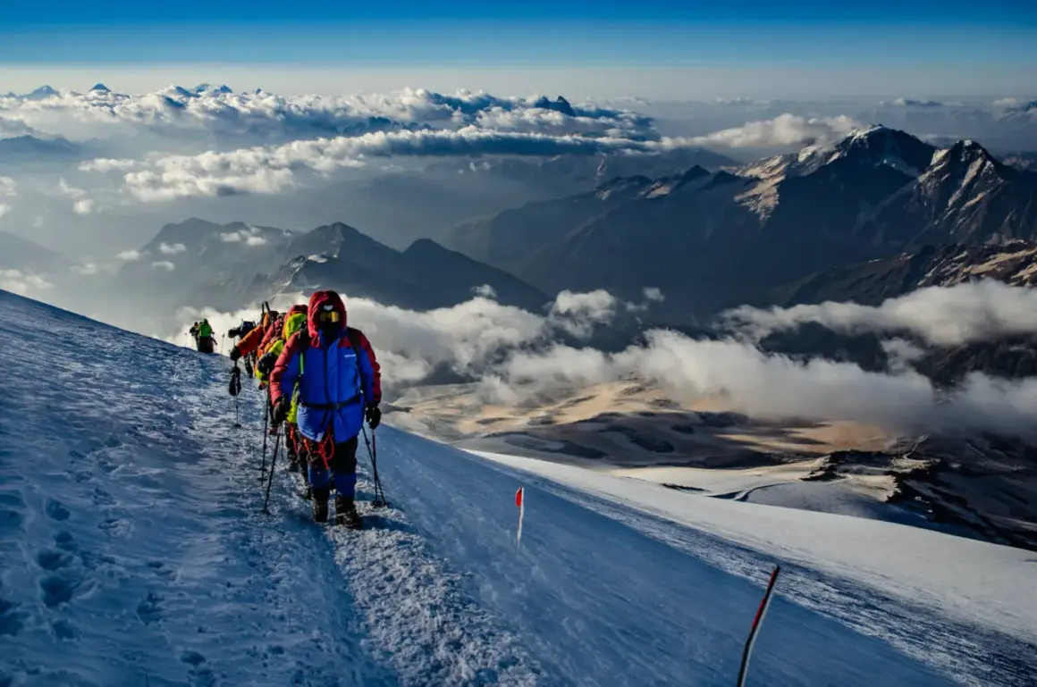 Grupa alpinistów wspina się po ośnieżonym zboczu, podziwiając majestatyczne góry z Elbrusem w tle. Chmury unoszą się poniżej, tworząc baśniowy krajobraz.