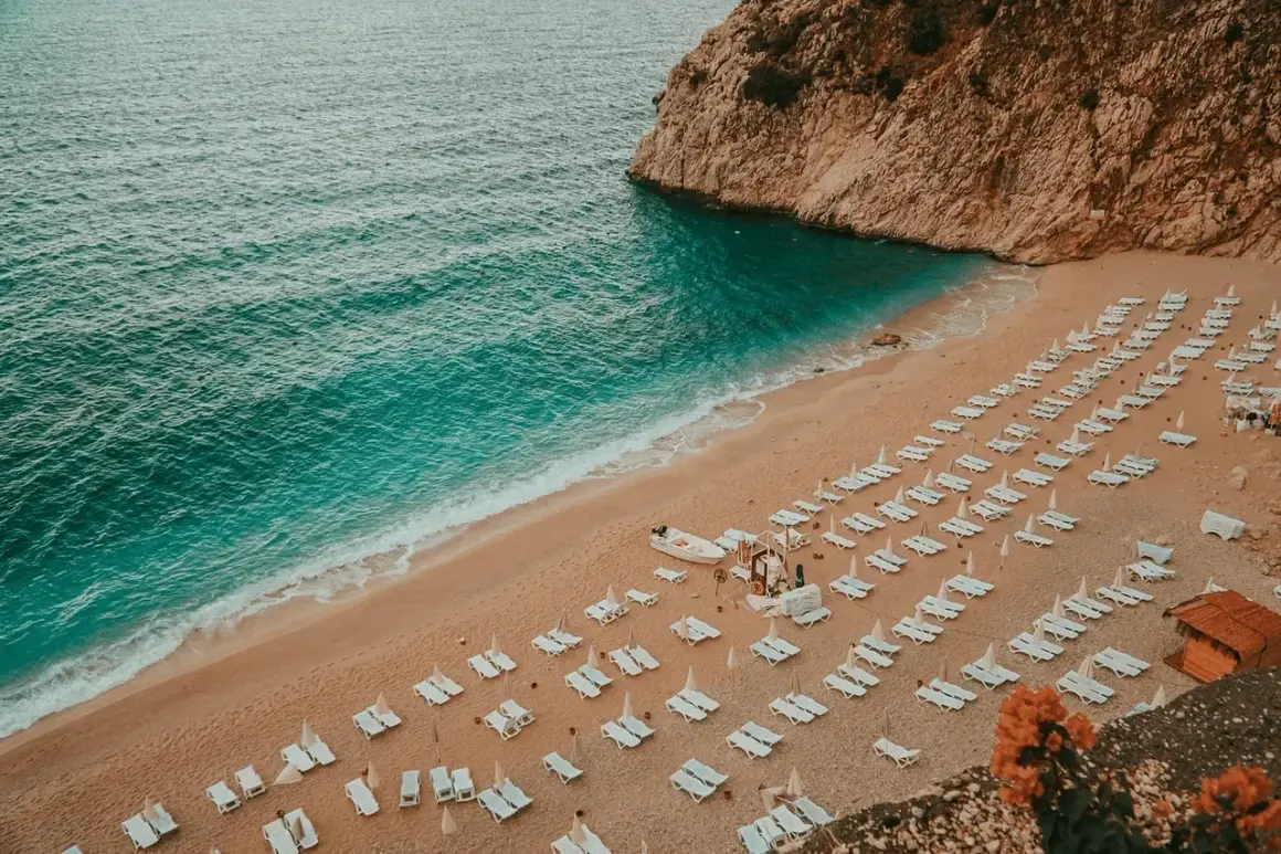 Plaża w Turcji, turkusowe morze, piaszczysty brzeg z rzędami leżaków i parasoli, skaliste wybrzeże.