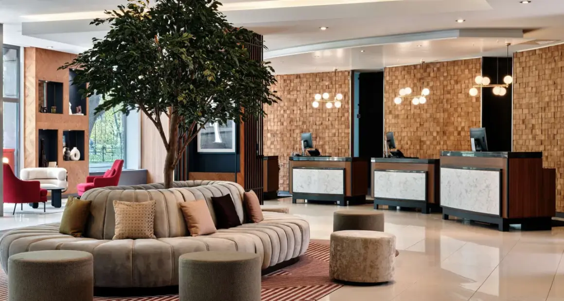 Eleganckie lobby hotelu Radisson Blu Hotel Krakow z okrągłą sofą, drzewem i recepcją z drewnianą ścianą.