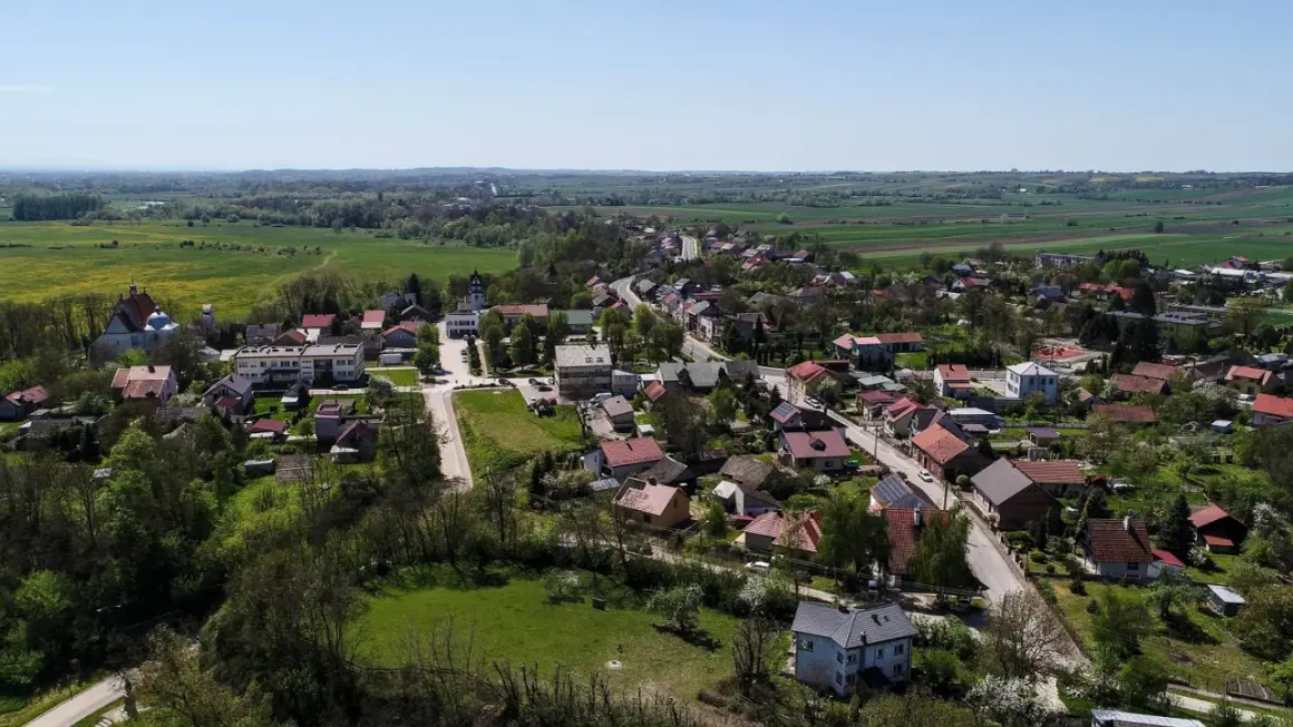 Widok z lotu ptaka na malowniczą wieś, która może pochwalić się mianem najmniejszego miasta w Polsce. Domy z czerwonymi dachami, kościół i zieleń pól tworzą sielski krajobraz.