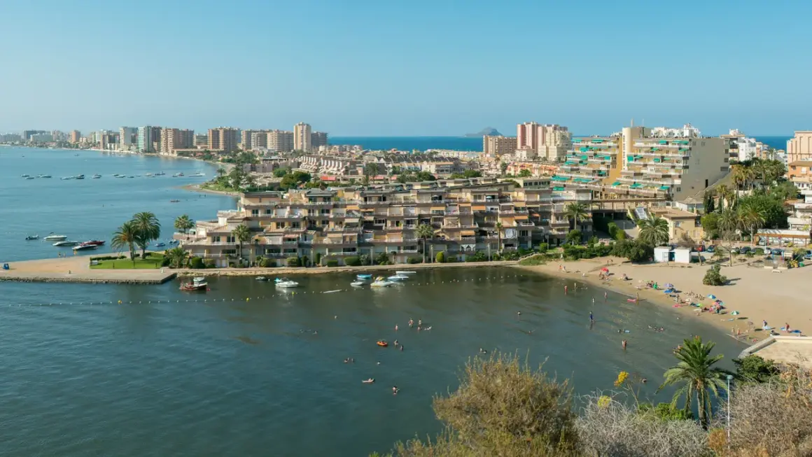 Widok na La Manga z plażą, budynkami mieszkalnymi i wodą pełną łodzi i kąpiących się ludzi.