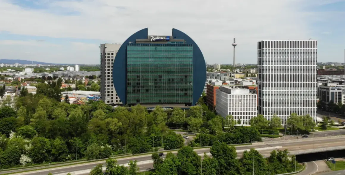 Charakterystyczny budynek Radisson Blu we Frankfurcie, otoczony zielenią, obok nowoczesnych wieżowców.