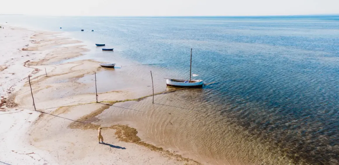 Spokojna plaża w Kuźnicy, gdzie kilka łodzi czeka na wodzie, a samotna postać spaceruje brzegiem.