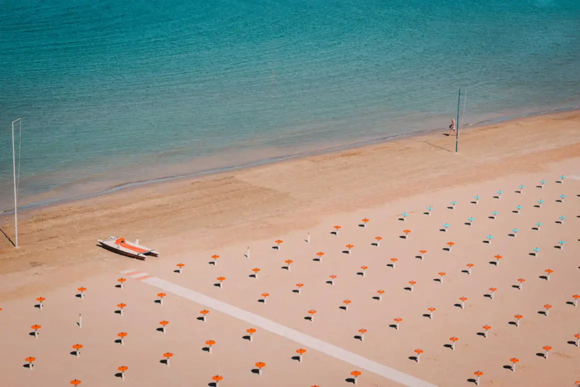 Pusta plaża w Rimini z rzędem pomarańczowych parasoli i małą łodzią. Woda jest turkusowa.
