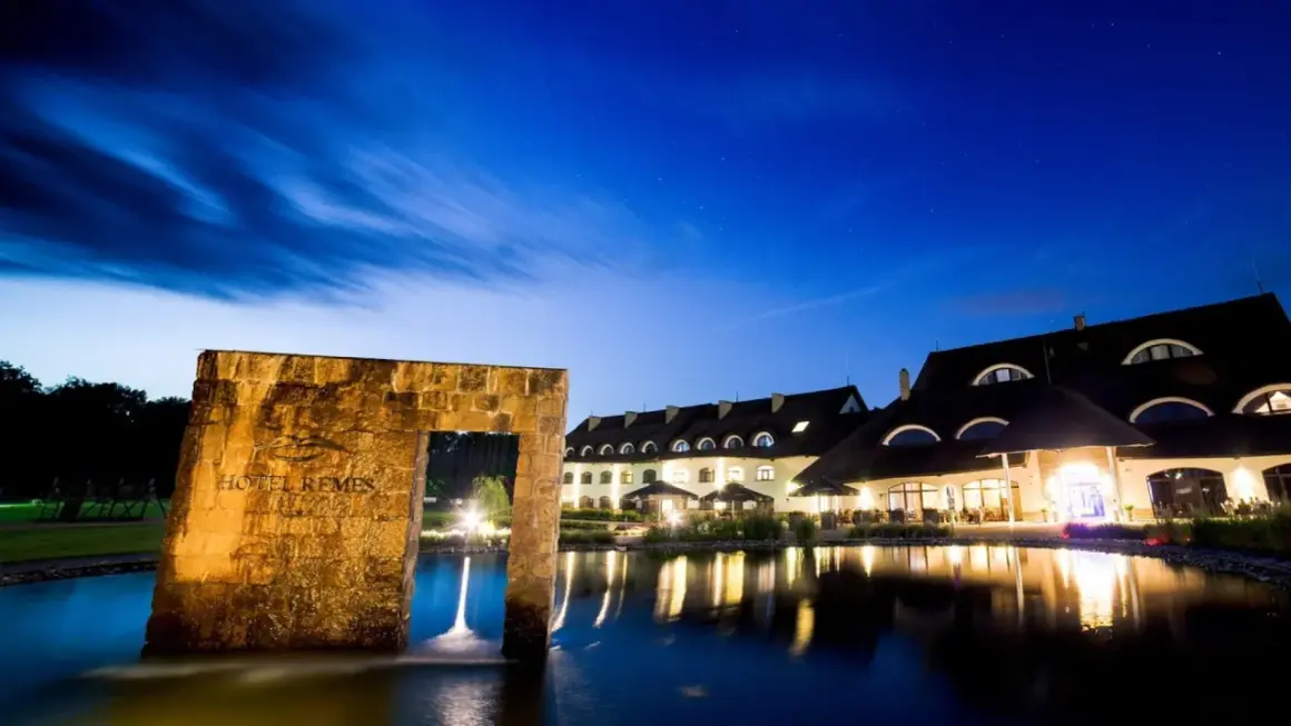 Hotel Remes Sport & Spa nocą, z kamiennym łukiem i stawem w tle.
