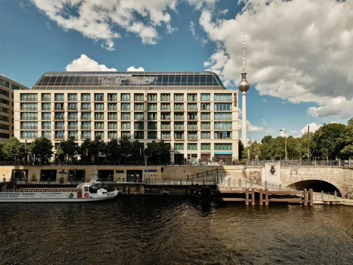Nowoczesny budynek hotelu Radisson Blu Berlin nad rzeką Sprewą, z widoczną w tle wieżą telewizyjną.