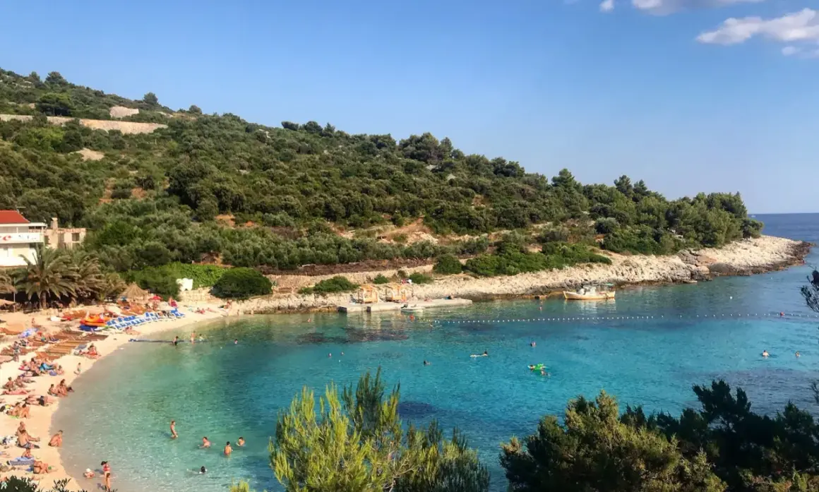Słoneczna plaża na wyspie Vis, z turkusową wodą, zielonymi wzgórzami i ludźmi odpoczywającymi na piasku.