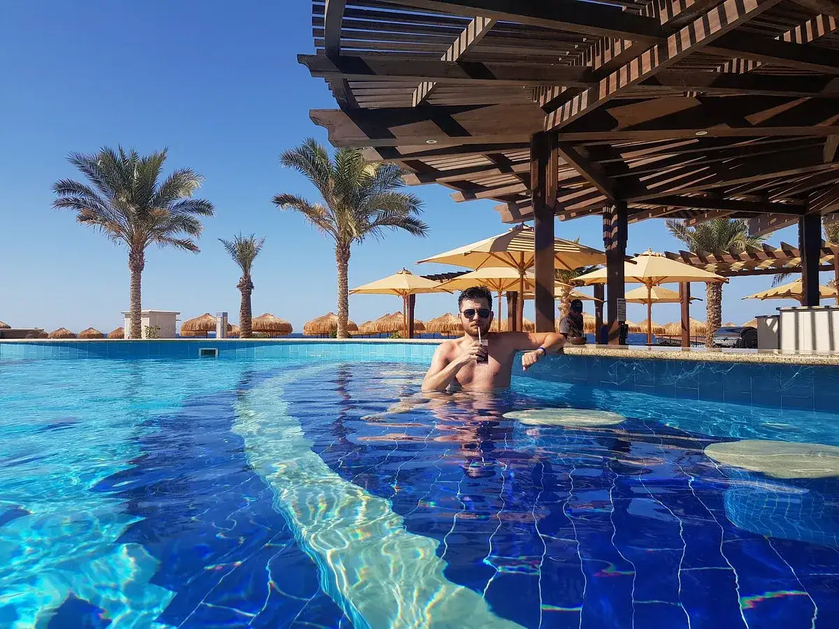 Mężczyzna z drinkiem w basenie przy barze w hotelu Lazuli Marsa Alam. Palmy i parasole na plaży w tle.
