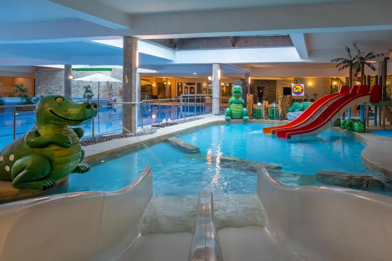 Wesoły hotel z aquaparkiem w górach zaprasza do zabawy! Dwa zielone dinozaury i zjeżdżalnie czekają na gości.