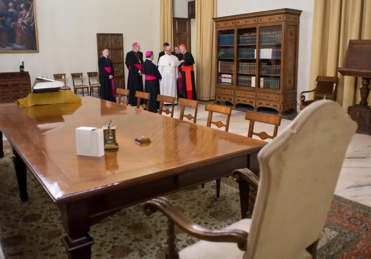 Papież Franciszek w otoczeniu duchownych w Domus Sanctae Marthae, przy dużym drewnianym stole.