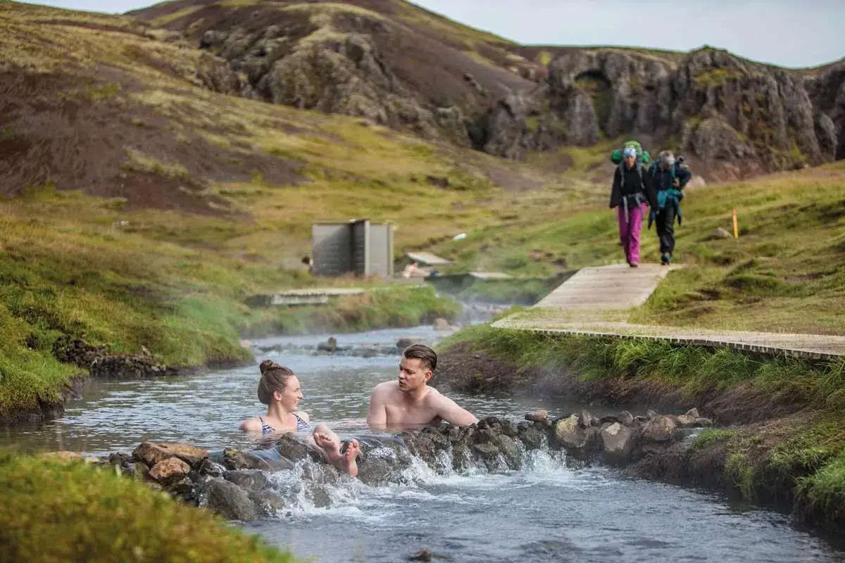 Para relaksu w gorących źródłach, niczym w blue lagoon, para zanurza się w strumieniu. W tle wędrowcy idą ścieżką.