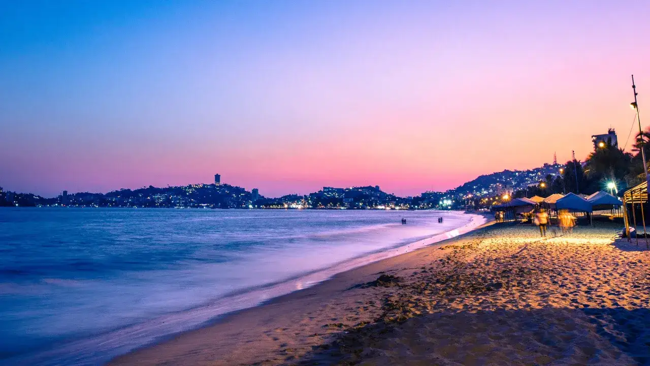 Zachód słońca nad Acapulco. Fale delikatnie obmywają piaszczystą plażę, a w oddali migoczą światła miasta na wzgórzach.
