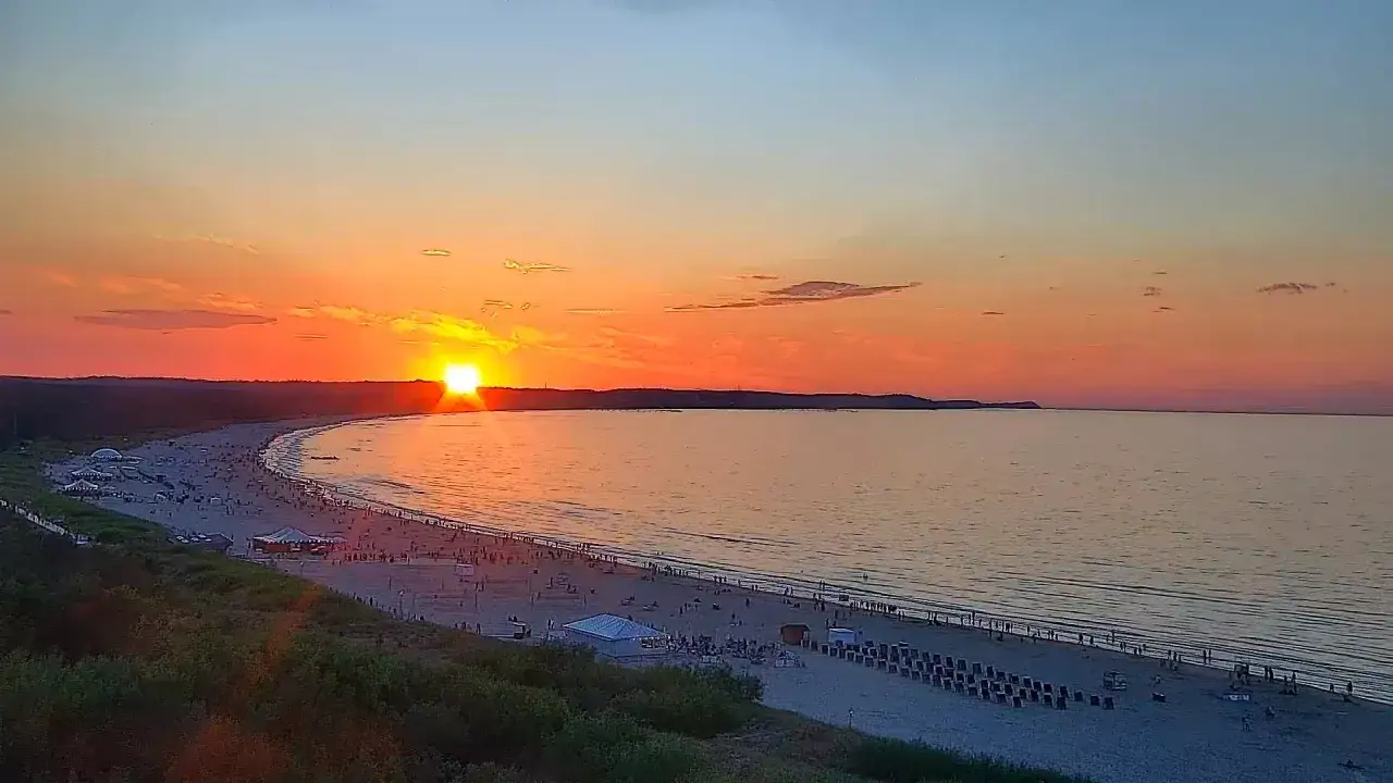 Piękny zachód słońca nad Bałtykiem w Świnoujściu. Słońce schodzi za horyzont, malując niebo odcieniami pomarańczy i różu. Plaża pełna ludzi cieszących się ostatnimi promieniami dnia.