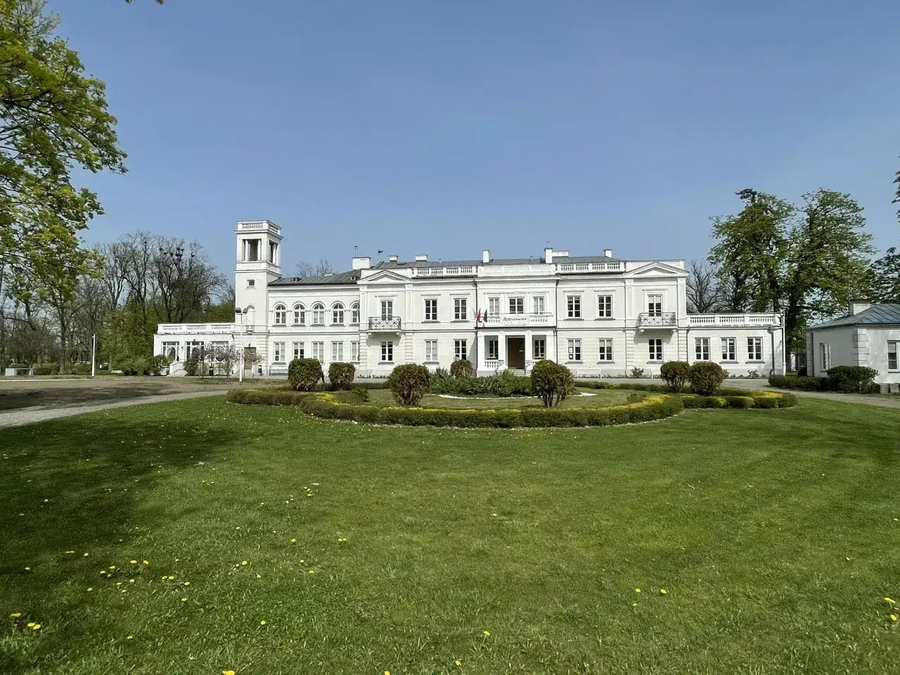 Pałac w Kiernozi, biała, okazała budowla z wieżą, otoczona zielonym ogrodem z klombami i drzewami.