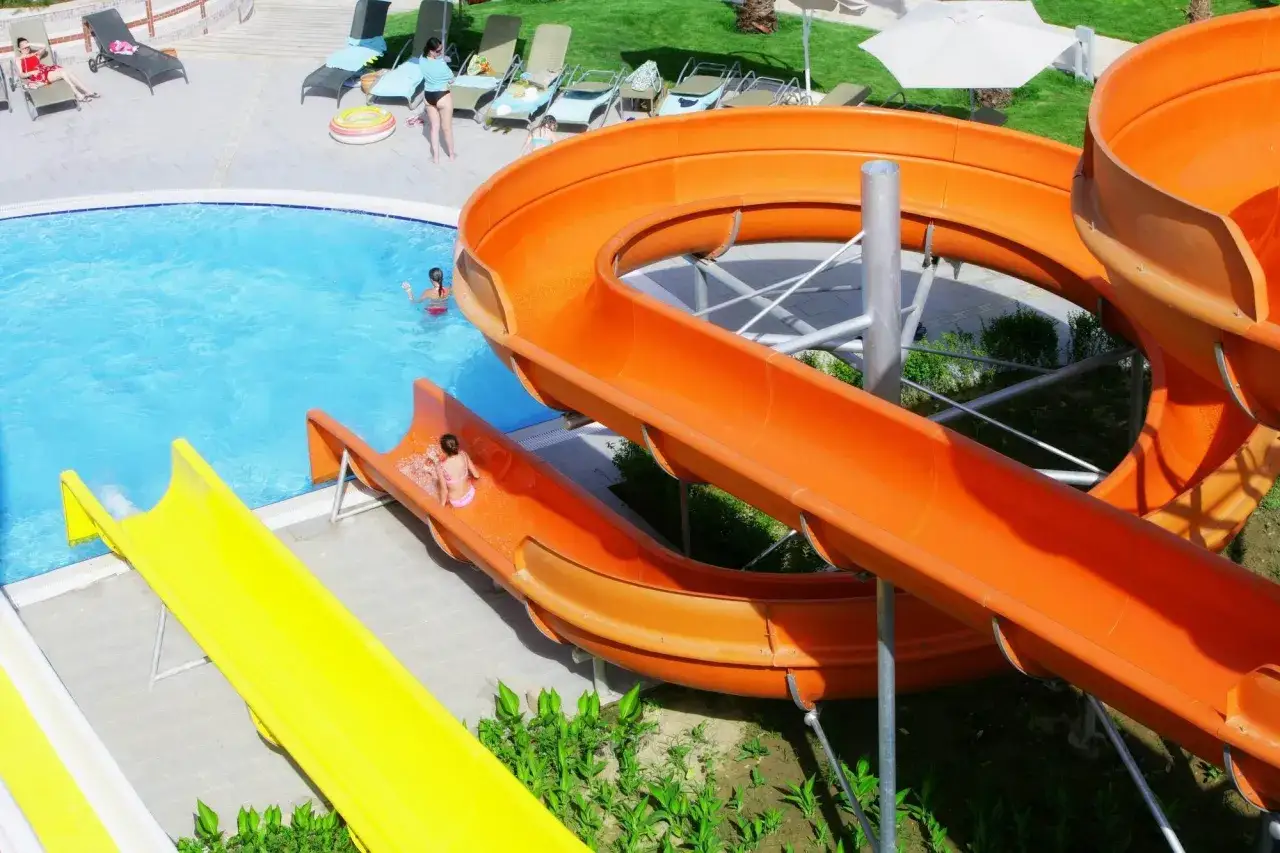 Dzieci zjeżdżają ze zjeżdżalni wodnych w basenie. Wiele osób odpoczywa na leżakach. Mukarnas spa resort opinie są pozytywne.