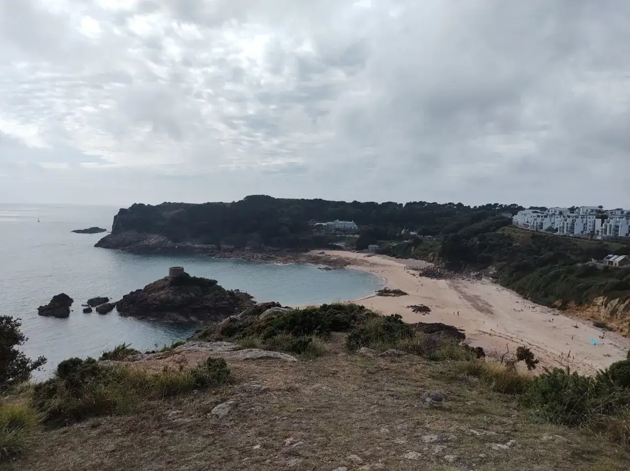 Widok na zatokę z piaszczystą plażą i wieżą na skale. W oddali widać zabudowania, a nad wszystkim chmury. To piękna wyspa Jersey.