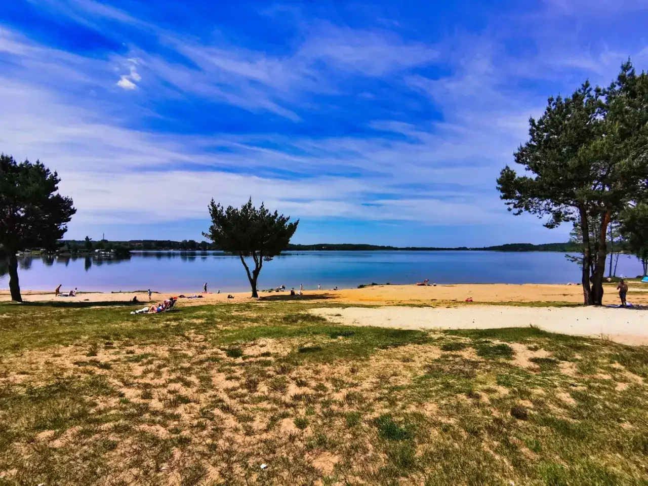 Pławniowice jezioro: piaszczysta plaża, błękitne niebo z chmurami, drzewa i ludzie odpoczywający nad wodą.